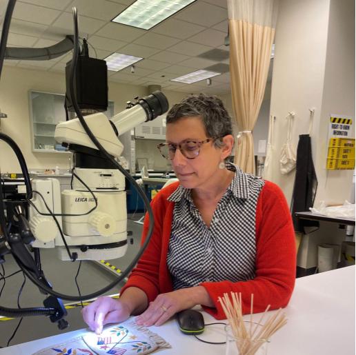 A white woman with short hair and glasses is looking at a historical object with a machine. The object is a leather pendant with painting of flowers and American flags.